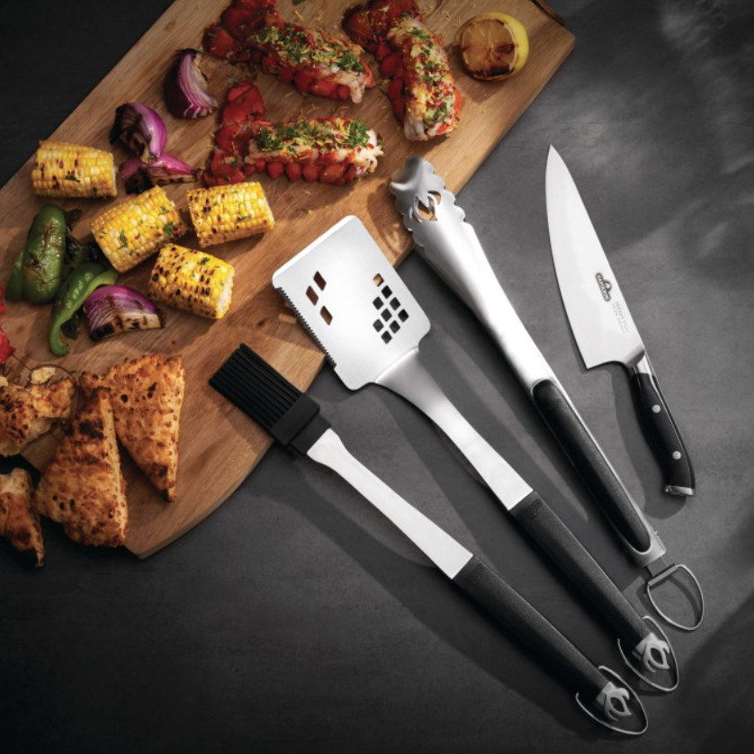 Grilled lobster tails, corn, peppers, and bread on a wooden cutting board alongside stainless steel grilling tools including tongs, spatula, basting brush, and chef’s knife on a dark surface.