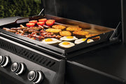 Grill with various foods cooking on a flat top, including eggs, tomatoes, and sausages.