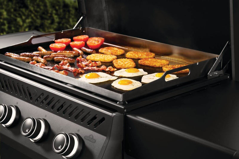 Grill with various foods cooking on a flat top, including eggs, tomatoes, and sausages.